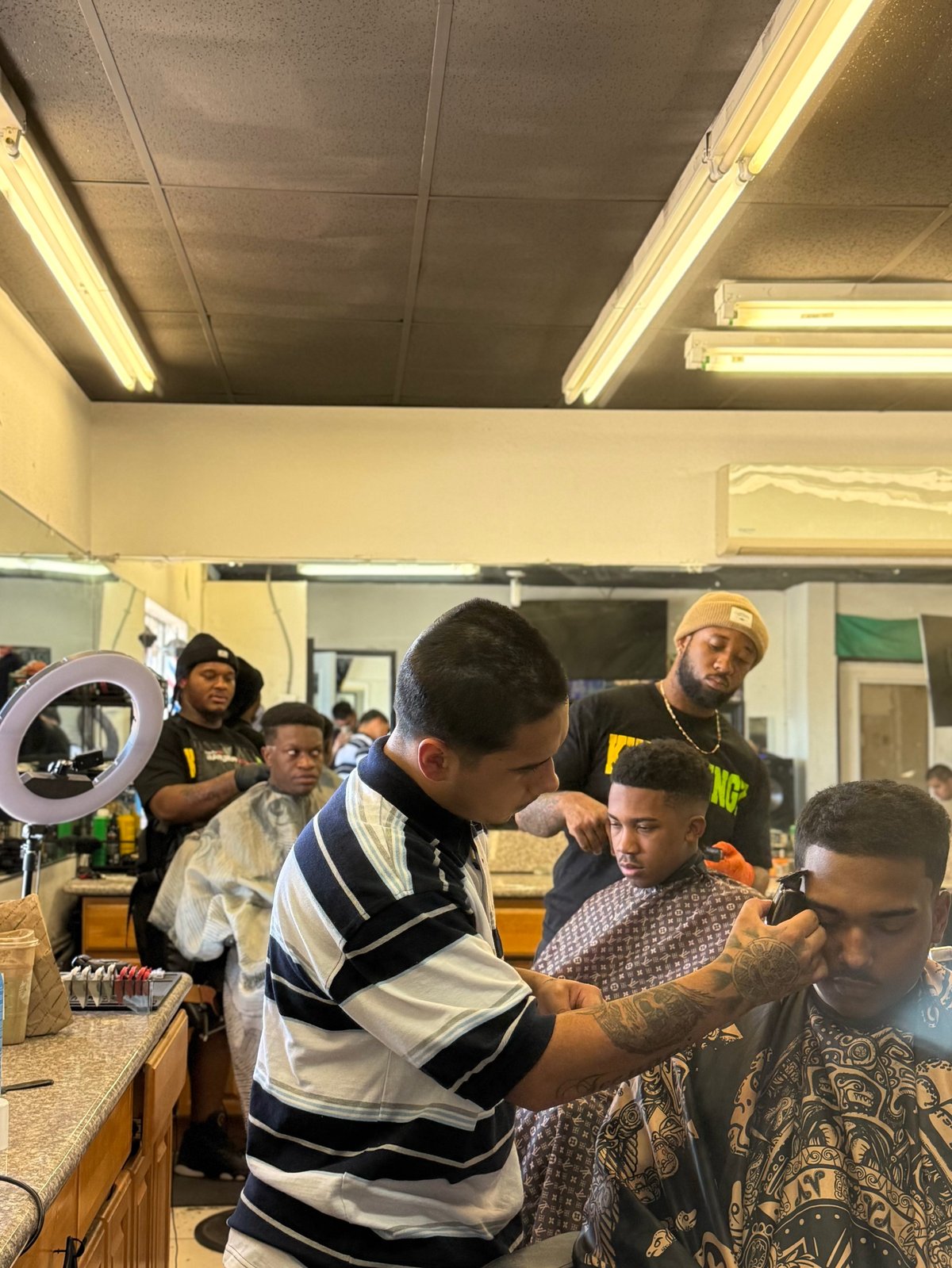 Barber cutting customers hair in a busy barbershop with multiple people waiting and observing the haircut
